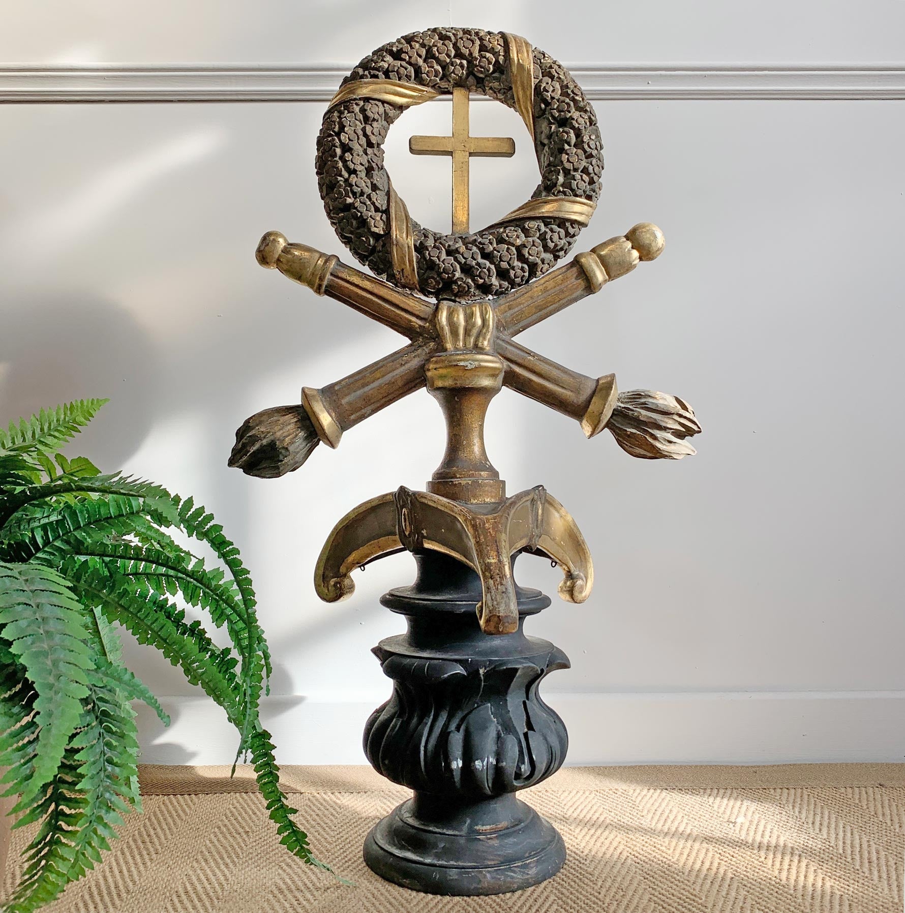 A decorative antique religious sculpture featuring a wreath with a cross, crossed swords, a celtic-style crest, and a dark pedestal base, styled indoors next to a green fern plant.