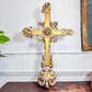 An ornate, antique gold and white carved wooden cross with a central circular medallion, displayed on a dark wooden surface against a white paneled wall.