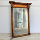 A large antique trumeau wall mirror with an ornate carved wooden frame, leaning against a white wall on a stone floor. The mirror reflects a green metal chair and interior setting.