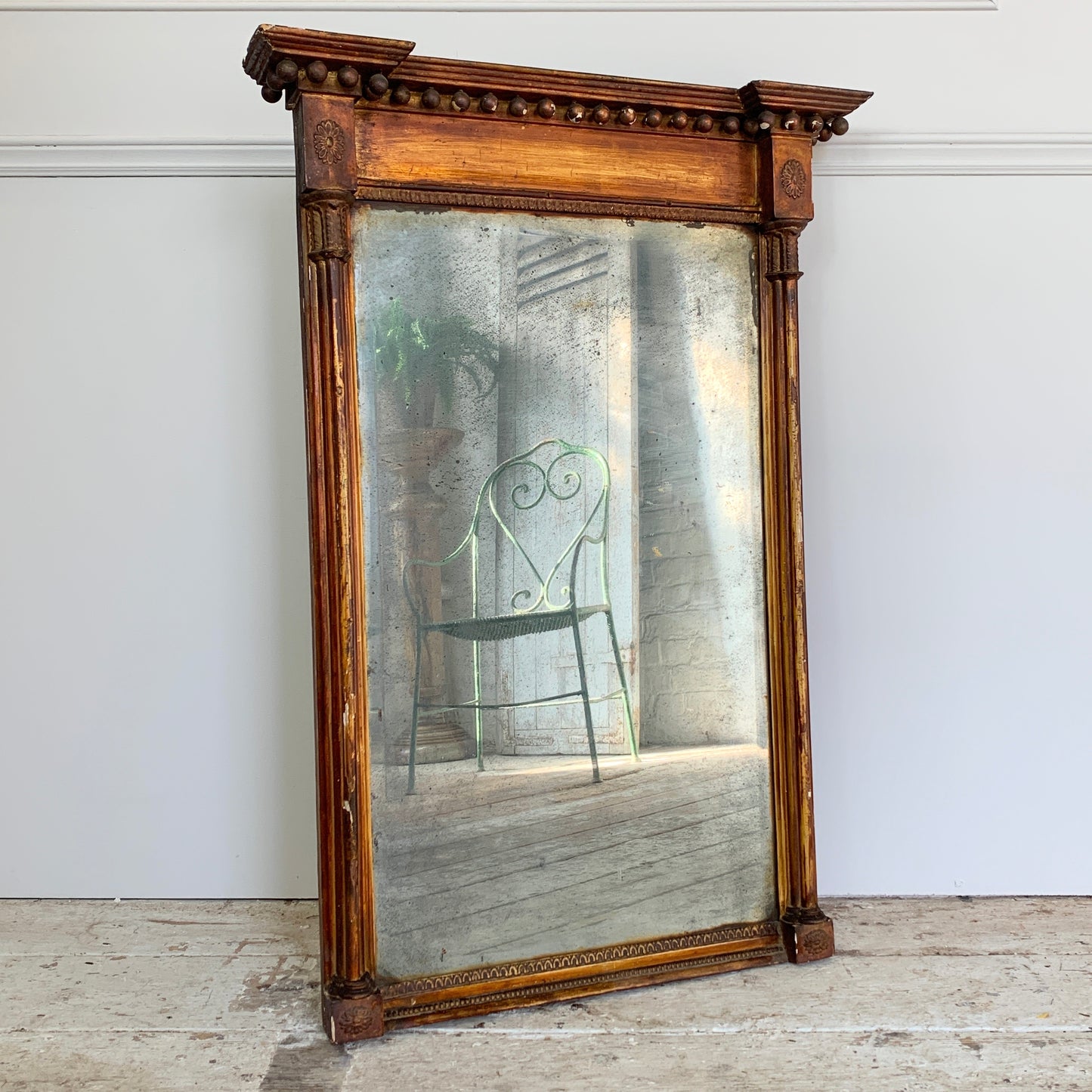 A large antique trumeau wall mirror with an ornate carved wooden frame, leaning against a white wall on a stone floor. The mirror reflects a green metal chair and interior setting.