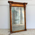 A large antique trumeau wall mirror with an ornate carved wooden frame, leaning against a white wall on a stone floor. The mirror reflects a green metal chair and interior setting.