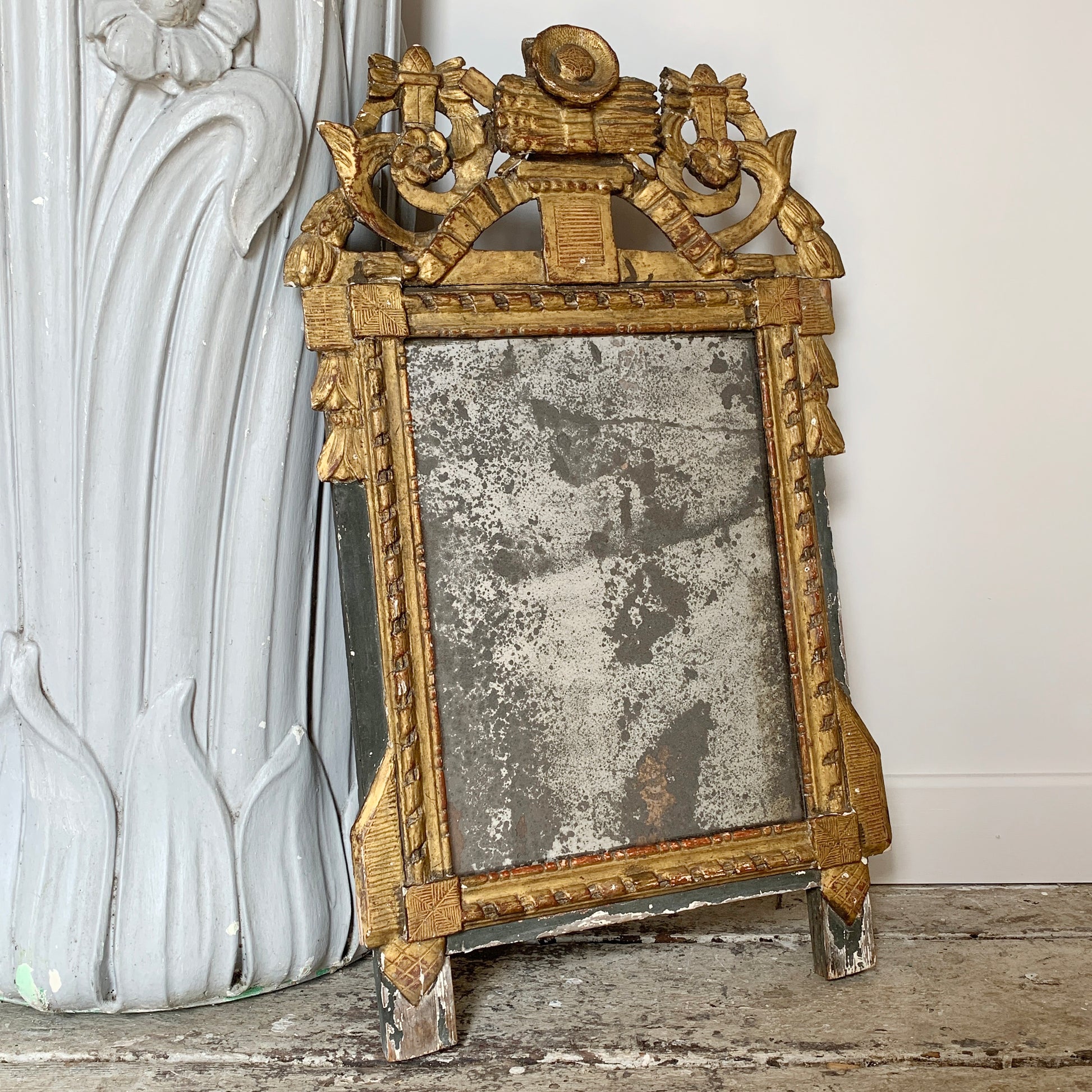 An ornate antique mirror with a distressed gold frame, leaning against a white decorative column on a stone floor.