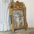 An ornate antique mirror with a distressed gold frame, leaning against a white decorative column on a stone floor.