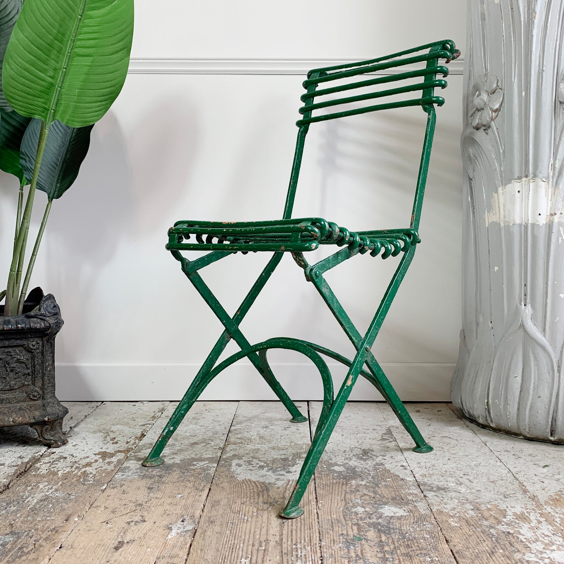 An antique green arras metal folding chair with a slatted back and seat, positioned on a rustic wooden floor against a white wall, flanked by a potted plant on the left and a distressed white decorative column on the right.