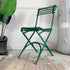An antique green arras metal folding chair with a slatted back and seat, positioned on a rustic wooden floor against a white wall, flanked by a potted plant on the left and a distressed white decorative column on the right.