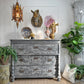 A distressed antique gray wooden three-drawer chest of drawers styled in a vintage room setting, decorated with a blue patterned plate, a horse head wall mount, a pink crystal cluster, and a vase, flanked by potted green plants.