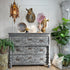 A distressed antique gray wooden three-drawer chest of drawers styled in a vintage room setting, decorated with a blue patterned plate, a horse head wall mount, a pink crystal cluster, and a vase, flanked by potted green plants.