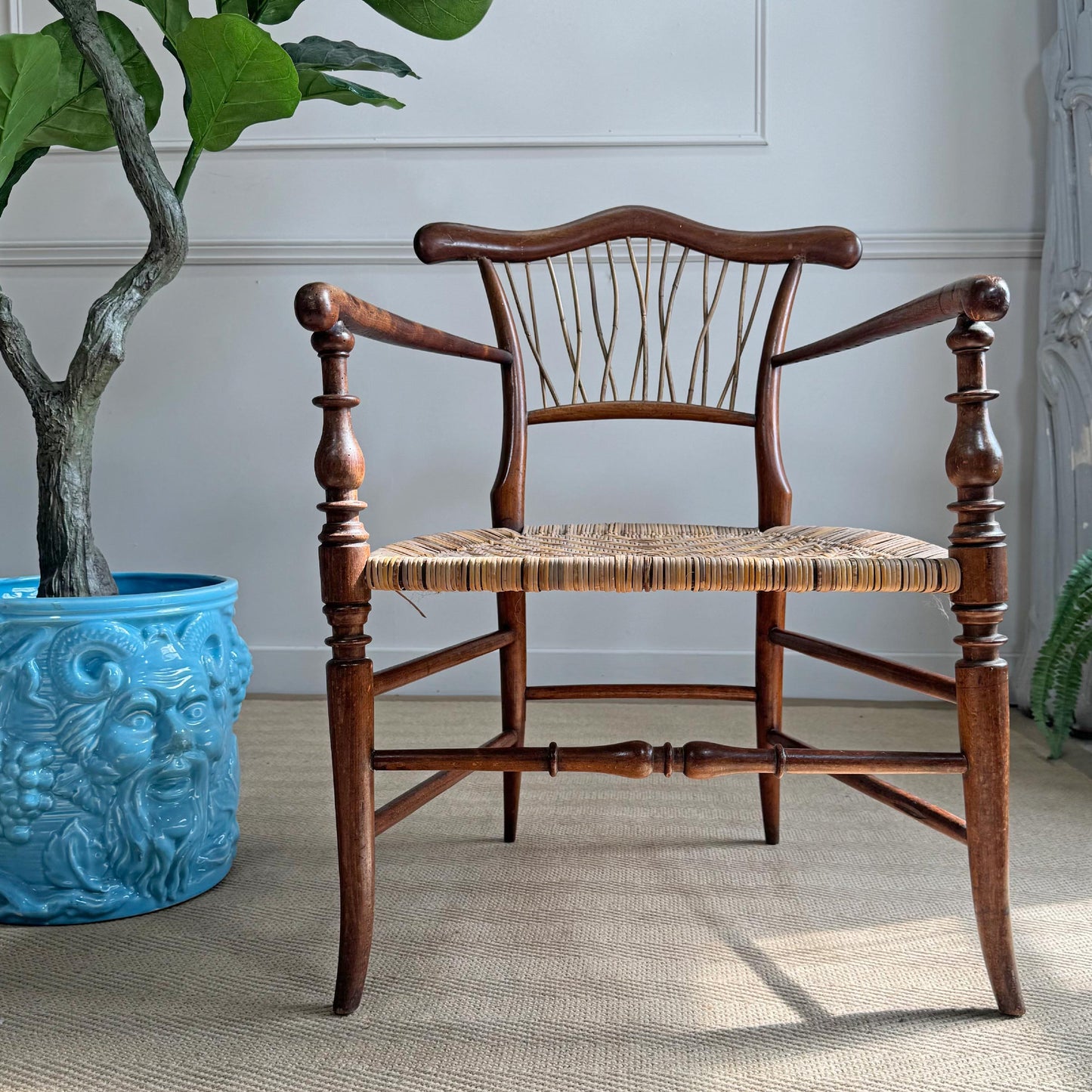 A dark brown antique wooden elbow armchair with a woven cane seat, positioned in front of a white paneled wall next to a large blue planter with a green plant.