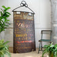 A rustic vintage-style metal wall sign with a black hanging chain, featuring faded multicolor advertising text including 'Pirelli'. The sign is styled in a home decor setting next to a green chair and potted ferns.