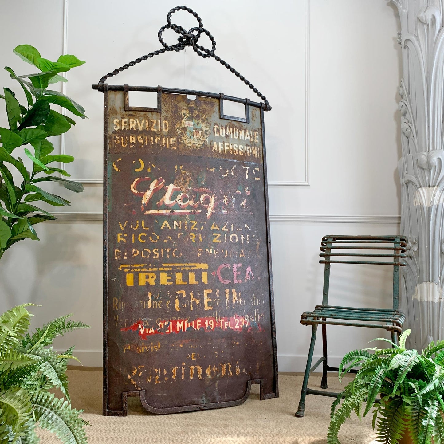 A rustic vintage-style metal wall sign with a black hanging chain, featuring faded multicolor advertising text including 'Pirelli'. The sign is styled in a home decor setting next to a green chair and potted ferns.