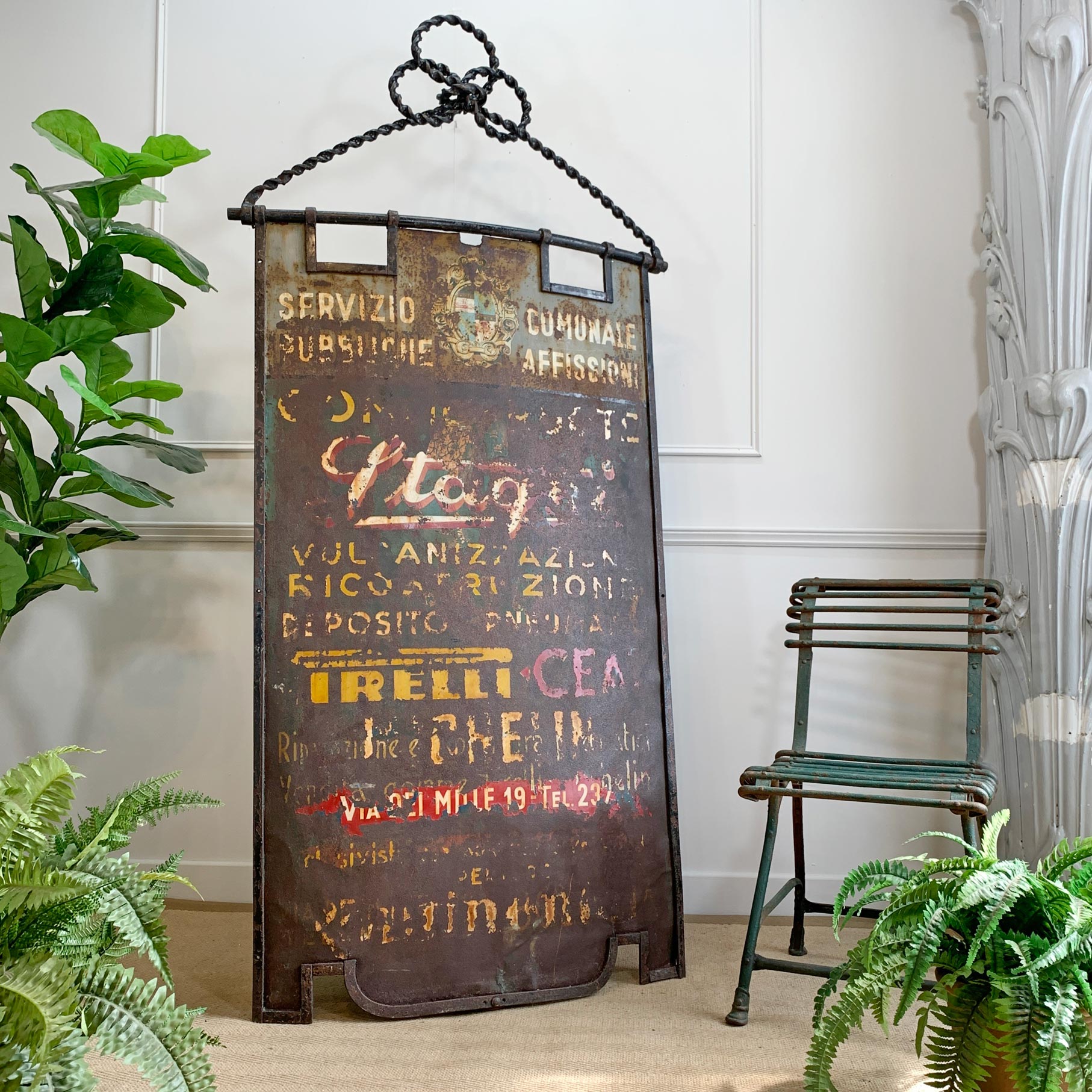 A rustic vintage-style metal wall sign with a black hanging chain, featuring faded multicolor advertising text including 'Pirelli'. The sign is styled in a home decor setting next to a green chair and potted ferns.