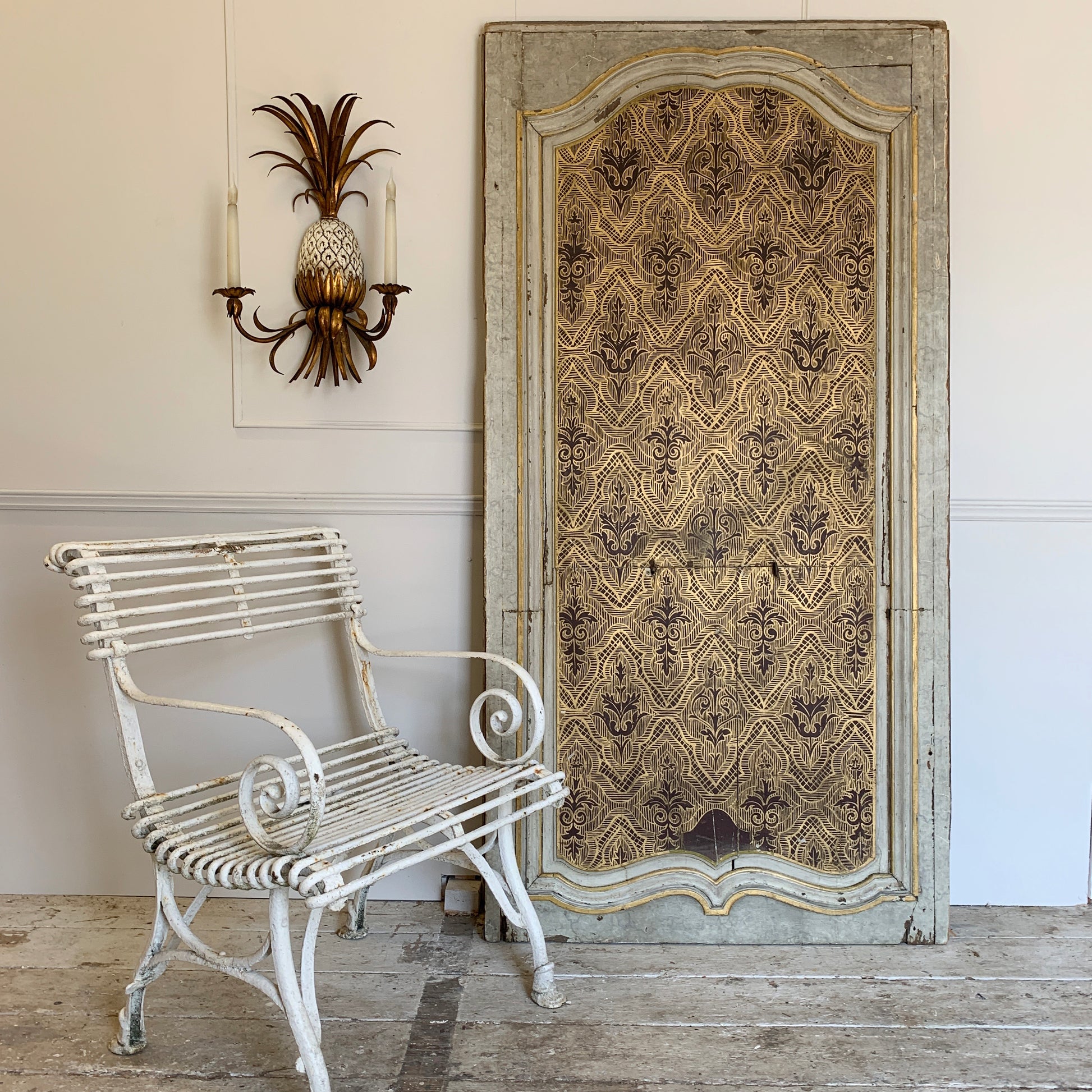 A tall, distressed vintage-style wooden panel with an ornate damask pattern in beige and brown, leaning against a white wall next to a white wrought-iron garden chair and a pineapple wall sconce.