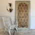 A tall, distressed vintage-style wooden panel with an ornate damask pattern in beige and brown, leaning against a white wall next to a white wrought-iron garden chair and a pineapple wall sconce.