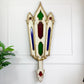 A decorative wall-mounted antique stained glass ornament with a distressed white and gold frame, featuring green, red, and blue glass panels, shown against a white paneled wall with a small plant at the side.