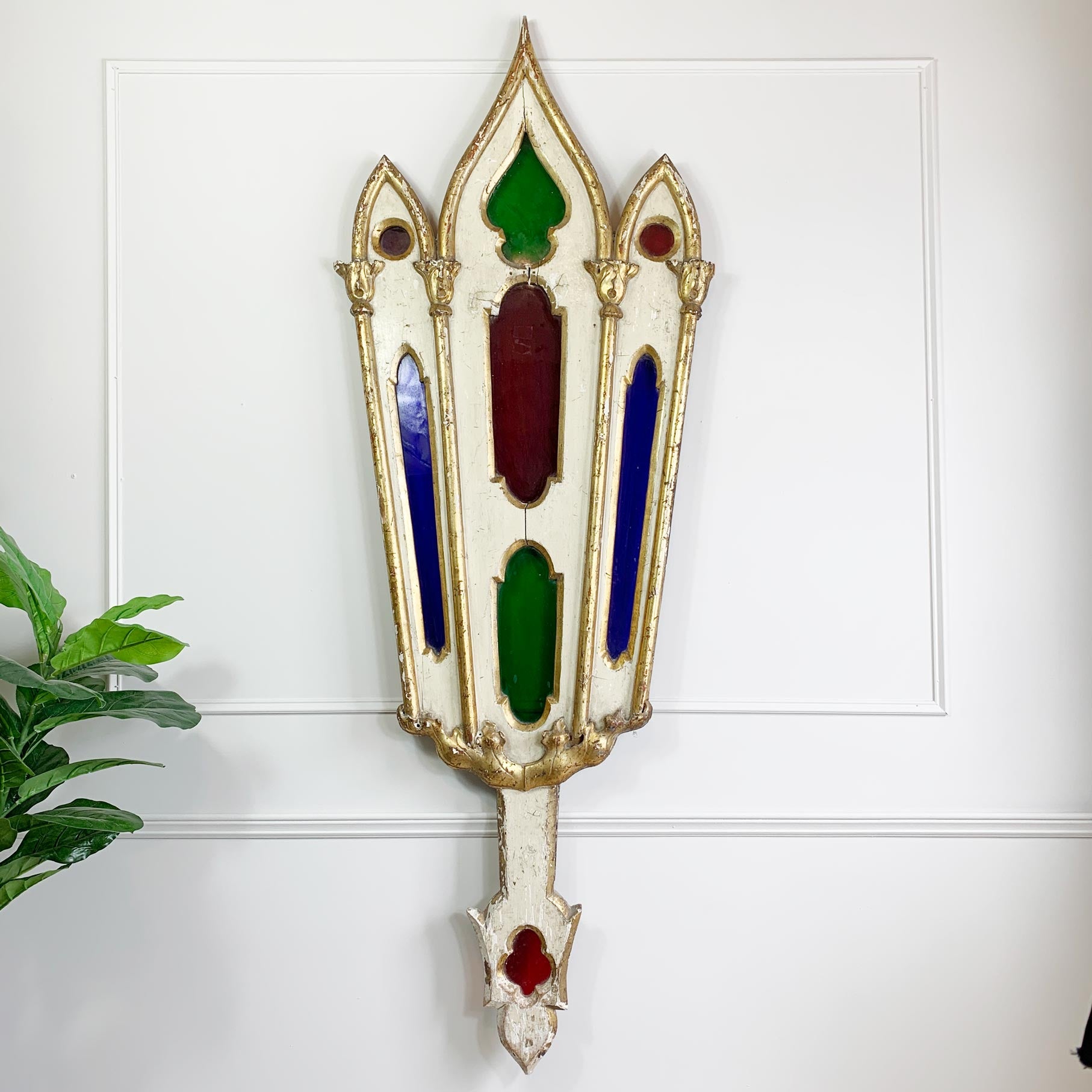 A decorative wall-mounted antique stained glass ornament with a distressed white and gold frame, featuring green, red, and blue glass panels, shown against a white paneled wall with a small plant at the side.