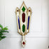 A decorative wall-mounted antique stained glass ornament with a distressed white and gold frame, featuring green, red, and blue glass panels, shown against a white paneled wall with a small plant at the side.