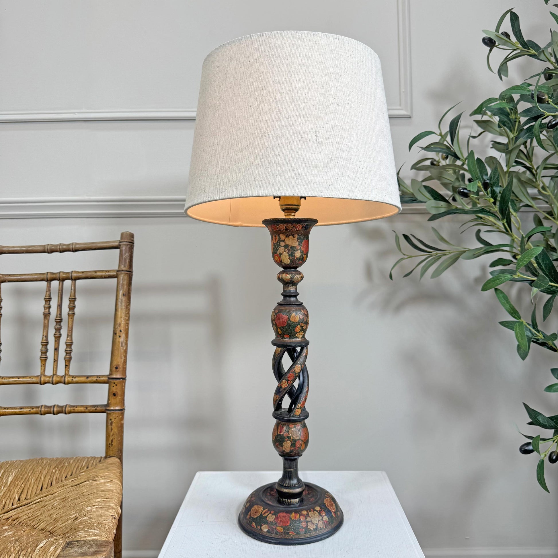 An antique decorative Kshmiri table lamp with a large off-white fabric drum shade and a dark carved wooden base featuring hand-painted floral details, set on a white pedestal against a light gray paneled wall with a wooden chair and green plant visible.