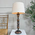 An antique decorative Kshmiri table lamp with a large off-white fabric drum shade and a dark carved wooden base featuring hand-painted floral details, set on a white pedestal against a light gray paneled wall with a wooden chair and green plant visible.