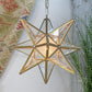 A six-pointed star-shaped pendant light with a gold metal frame and clear glass panels, glowing with a warm light. It hangs against an indoor background with a patterned textile and greenery visible.