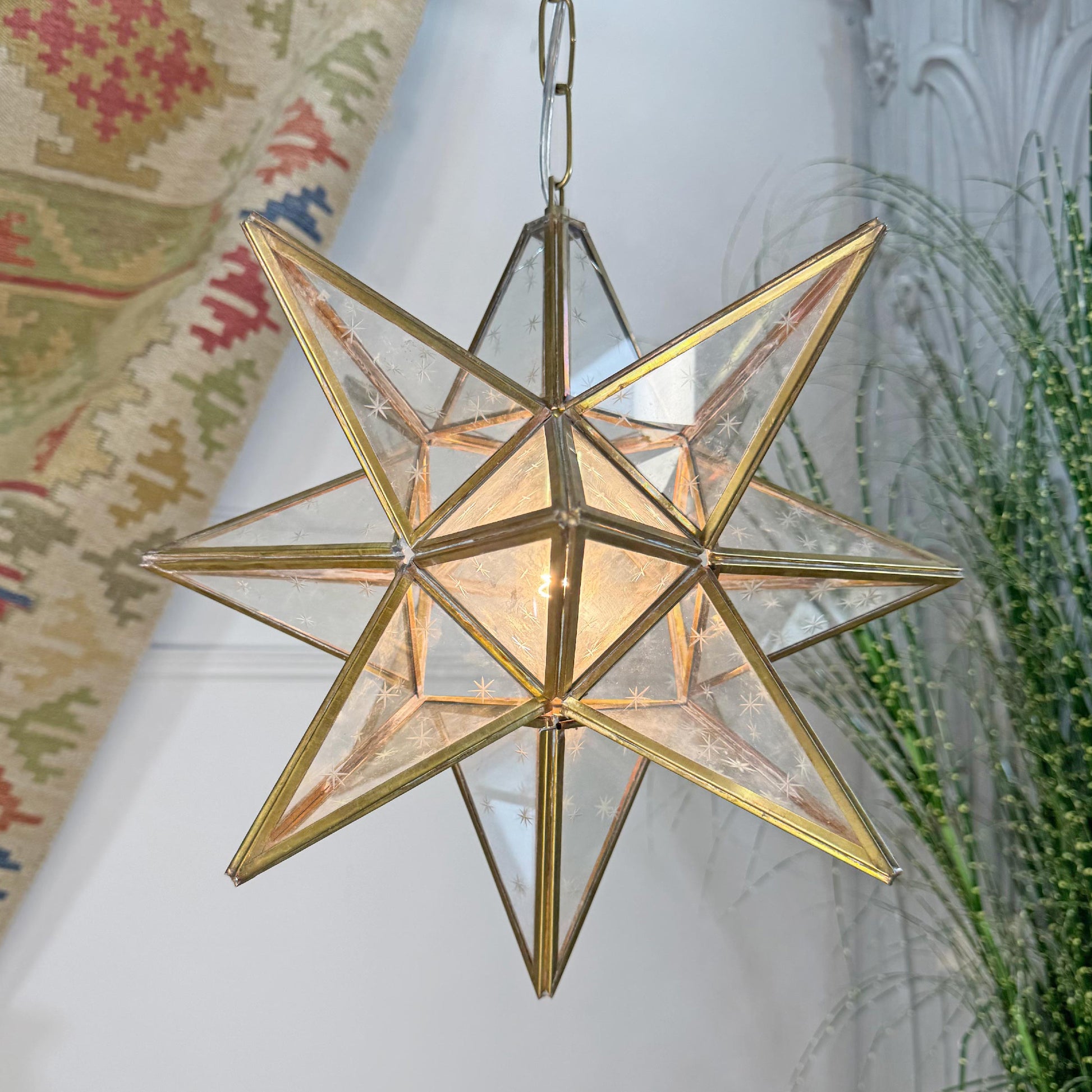 A six-pointed star-shaped pendant light with a gold metal frame and clear glass panels, glowing with a warm light. It hangs against an indoor background with a patterned textile and greenery visible.