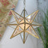 A six-pointed star-shaped pendant light with a gold metal frame and clear glass panels, glowing with a warm light. It hangs against an indoor background with a patterned textile and greenery visible.