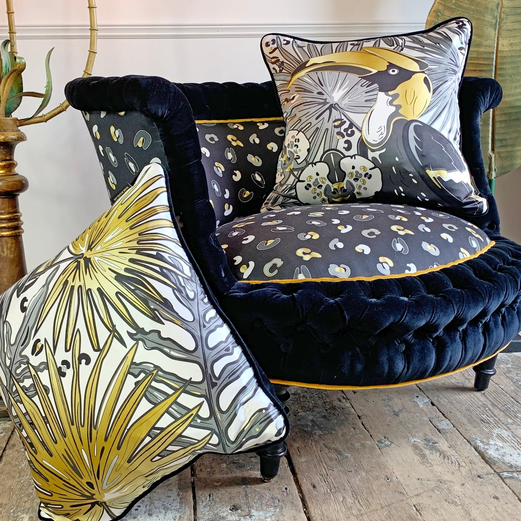 A buttoned velvet antique Marquise armchair upholstered in black velvet and leopard print, styled with three decorative throw pillows featuring a gold, gray, and cream tropical leaf and toucan print, set on a rustic wooden floor.
