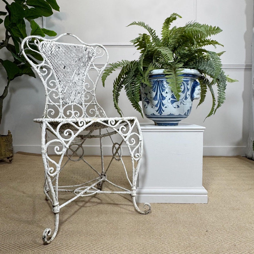 A distressed white wrought iron garden chair is styled next to a large Nevers French blue and white ceramic planter with a green fern, set on a beige jute rug against a white paneled wall.