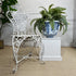 A distressed white wrought iron garden chair is styled next to a large Nevers French blue and white ceramic planter with a green fern, set on a beige jute rug against a white paneled wall.
