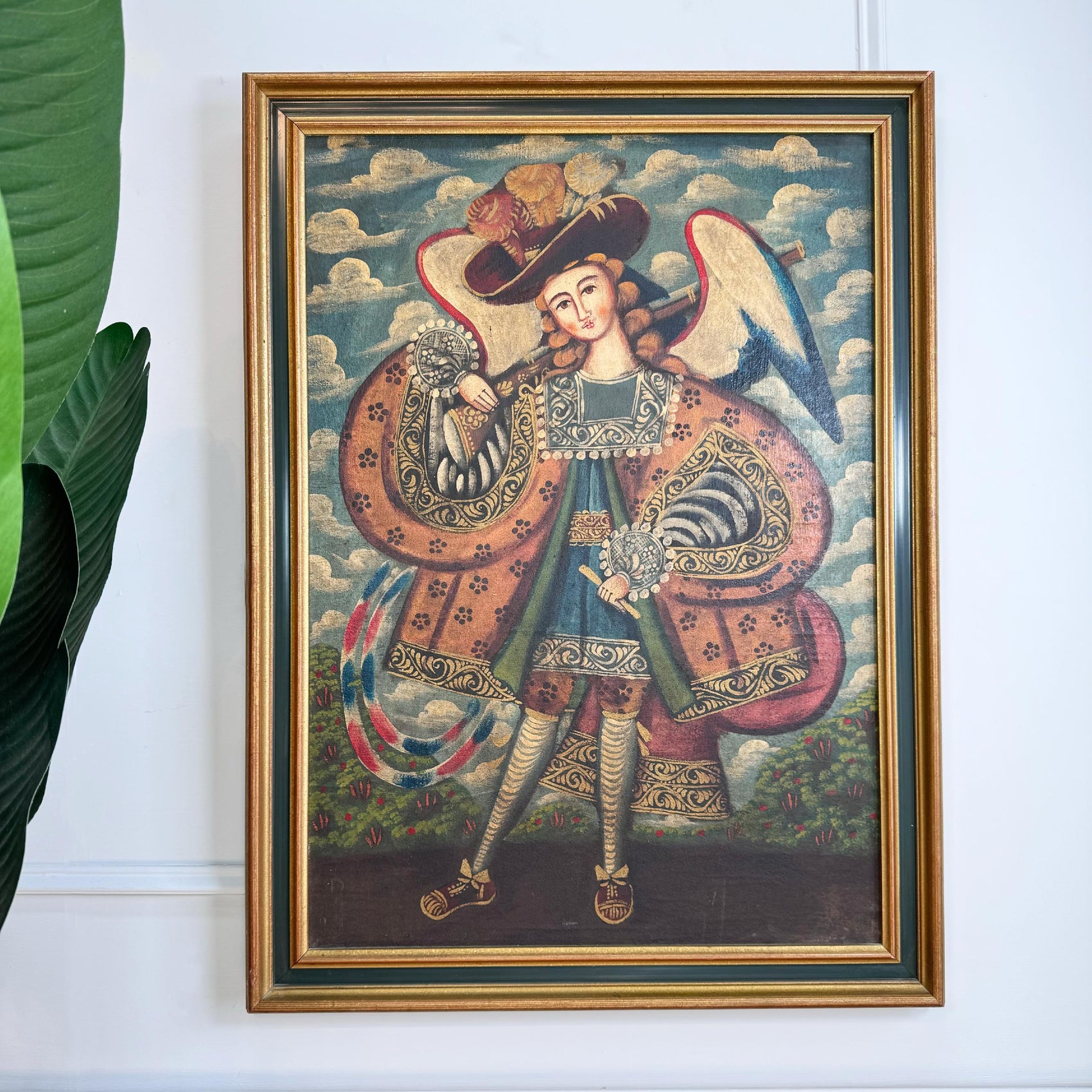 A framed antique Cuzco painting of a Archangel Raphael shown hanging on a white wall next to a green plant leaf. The painting features a child figure in ornate robes and a hat against a cloudy sky background, enclosed in a gold frame.