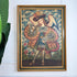 A framed antique Cuzco painting of a Archangel Raphael shown hanging on a white wall next to a green plant leaf. The painting features a child figure in ornate robes and a hat against a cloudy sky background, enclosed in a gold frame.