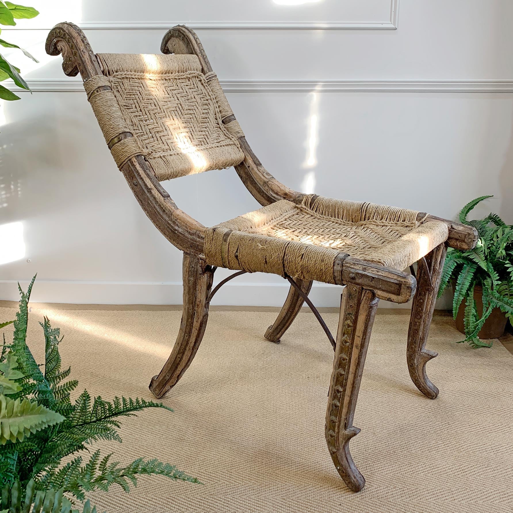A rustic wooden and woven rattan accent chair with curved legs, shown in a styled room setting with potted ferns and a neutral wall background.