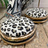 Two round velvet ottomans with leopard-print upholstery and distressed gold and black painted wood bases, styled on a rustic wooden floor with a fringed textile in the background.