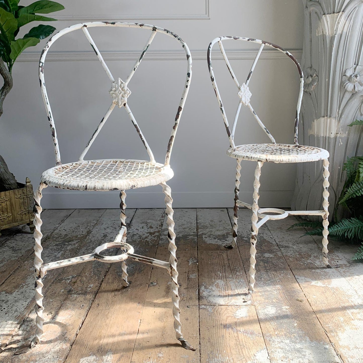 A pair of antique Regency metal chairs with distressed white paint and woven seats, placed on a rustic wooden floor against a light paneled wall.