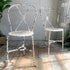 A pair of antique Regency metal chairs with distressed white paint and woven seats, placed on a rustic wooden floor against a light paneled wall.