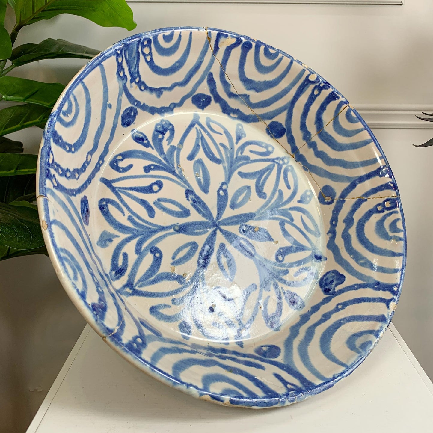 A large round ceramic antique Spanish bowl with a blue and white hand-painted Moroccan style pattern, displayed on a white surface with a green plant leaf in the background.