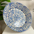 A large round ceramic antique Spanish bowl with a blue and white hand-painted Moroccan style pattern, displayed on a white surface with a green plant leaf in the background.