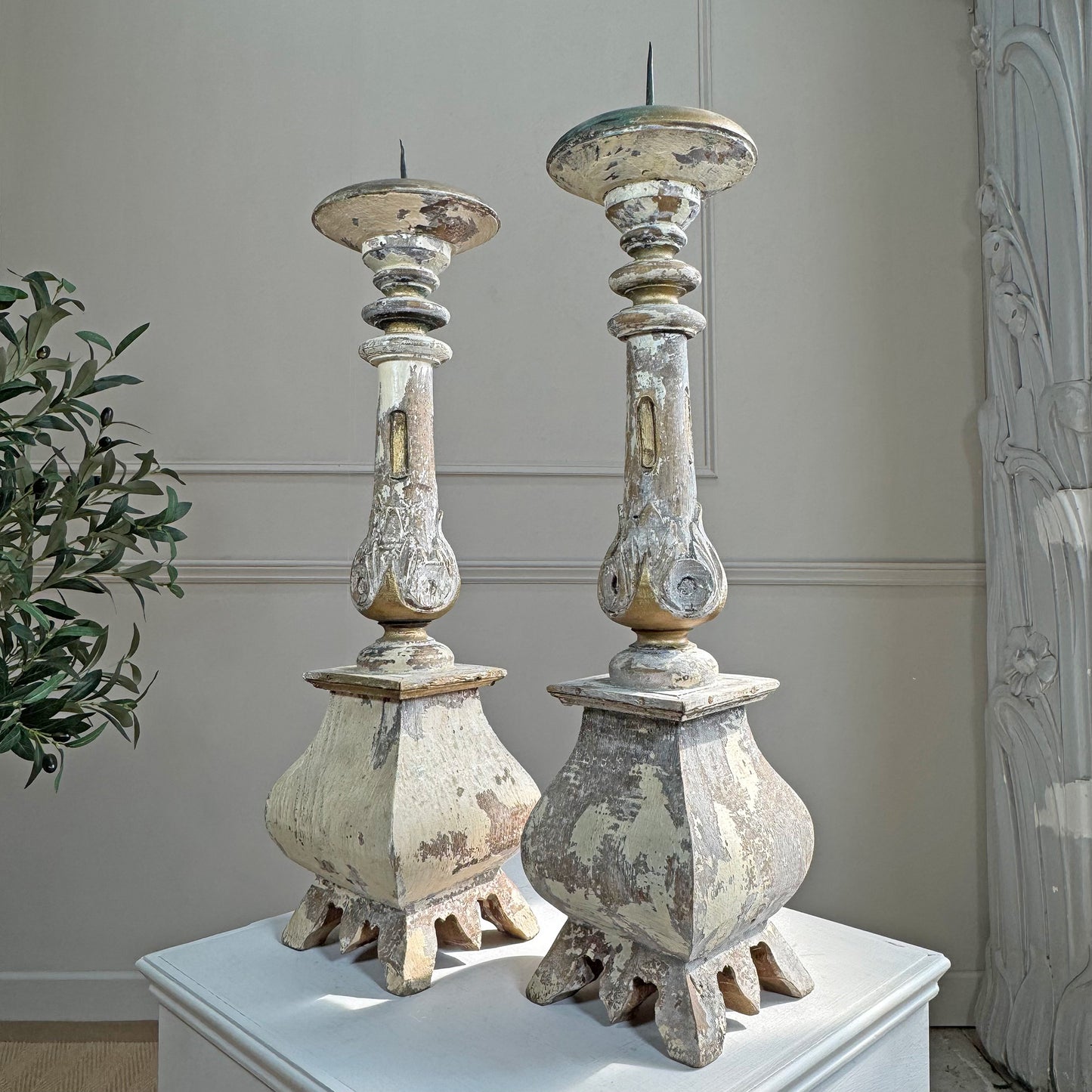 A pair of distressed, vintage-style candlestick holders with a weathered off-white and beige finish, displayed on a white pedestal in an indoor setting.
