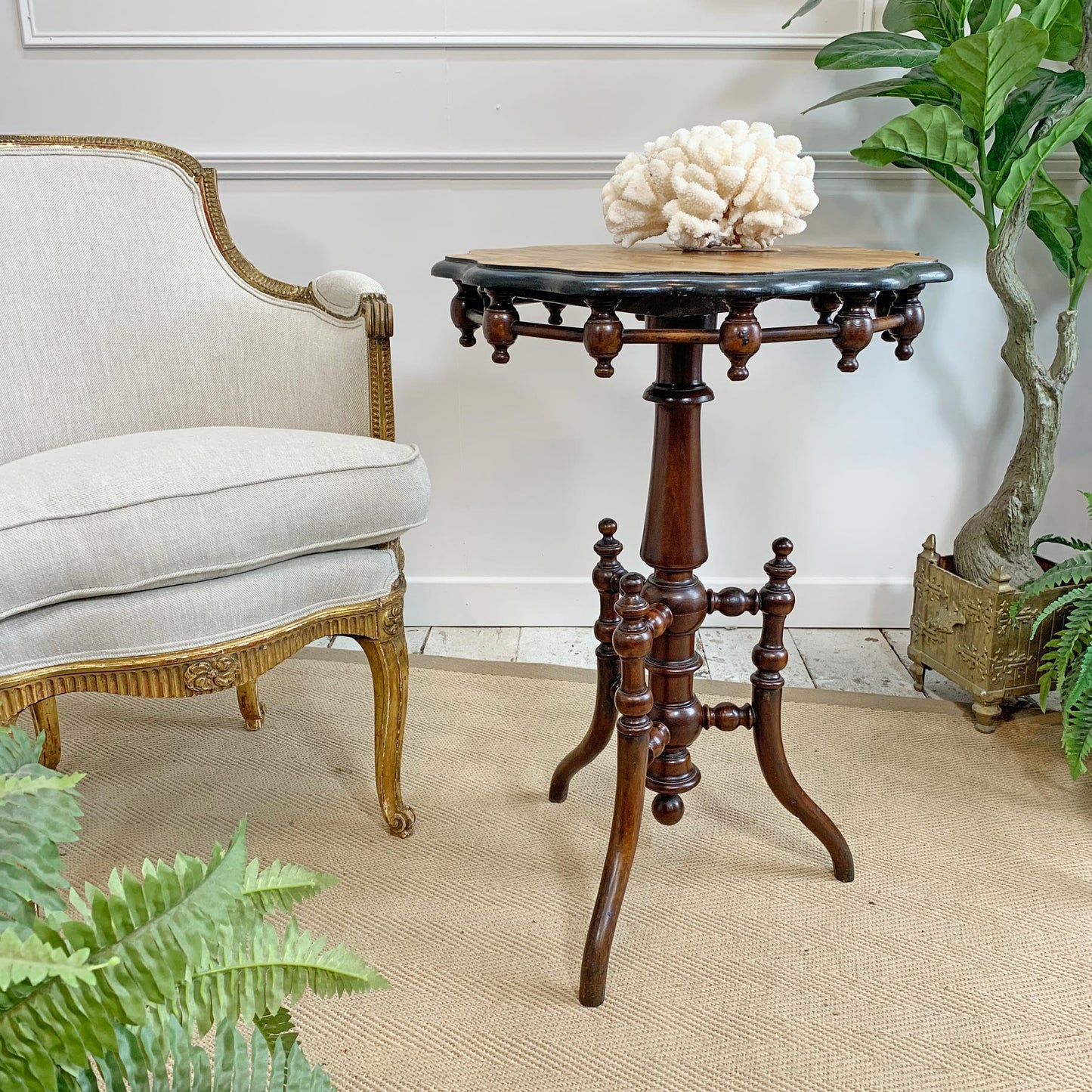 A small round antique swedish wooden pedestal side table with ornate turned legs and a dark carved apron, styled in a room next to a cream upholstered armchair with a coral sculpture on top.s