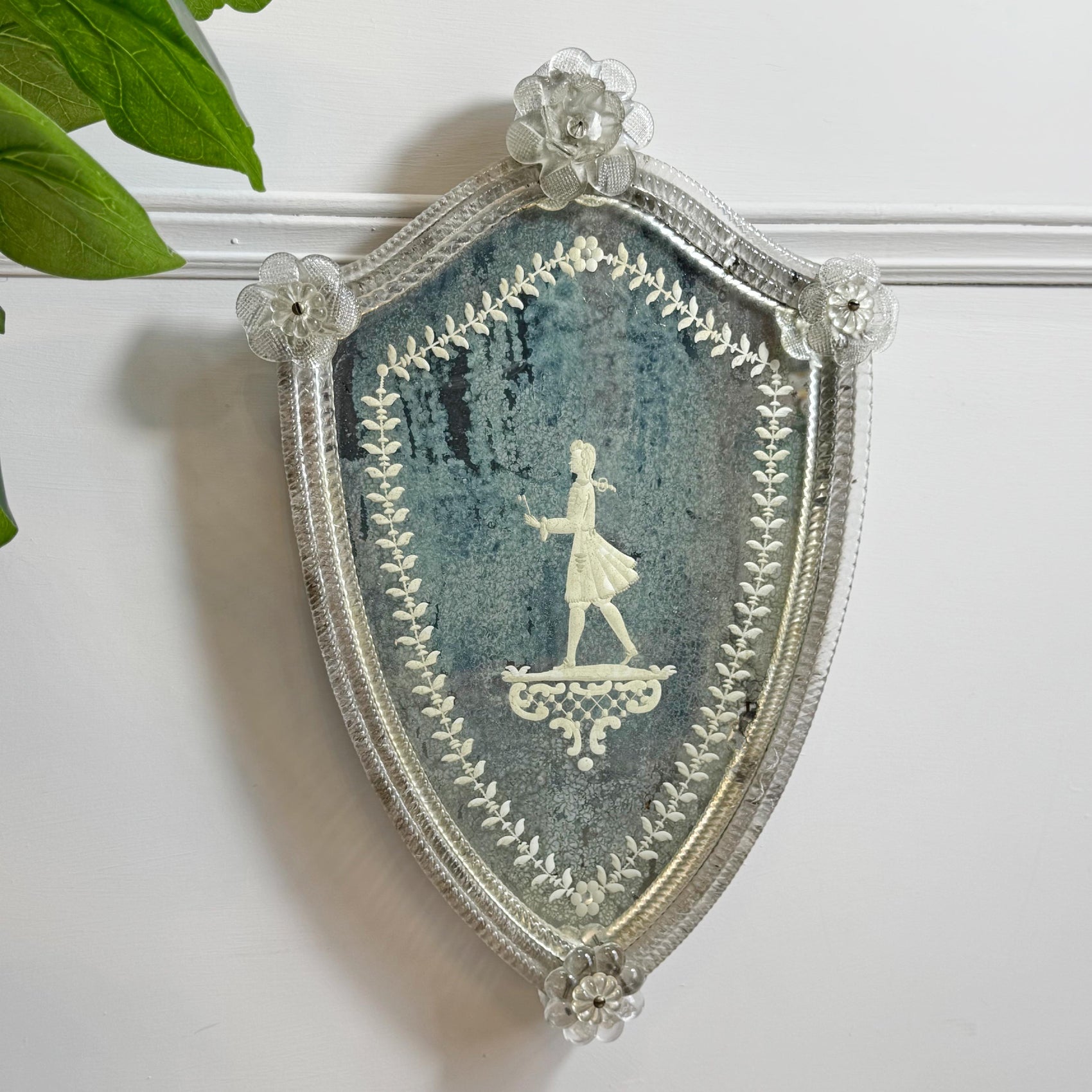 A decorative shield-shaped murano venetian mirror featuring a foxed mirror plate with silhouette of a man standing on a pedestal, surrounded by a silver-toned ornate border and floral accents at the corners, mounted on a white wall with a green plant leaf entering from the top left.