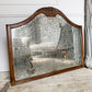A large antique wall mirror with an ornate carved wooden frame and a heavily textured, speckled mirrored glass surface, leaning against a white paneled wall on a rustic wooden floor.