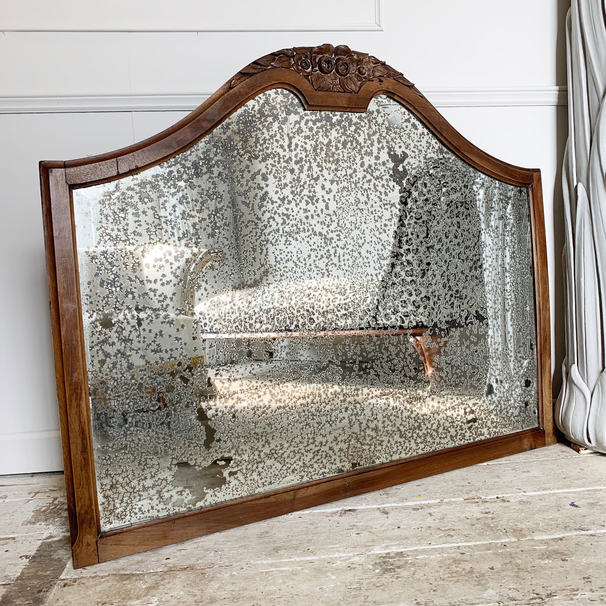 A large antique wall mirror with an ornate carved wooden frame and a heavily textured, speckled mirrored glass surface, leaning against a white paneled wall on a rustic wooden floor.