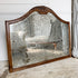A large antique wall mirror with an ornate carved wooden frame and a heavily textured, speckled mirrored glass surface, leaning against a white paneled wall on a rustic wooden floor.