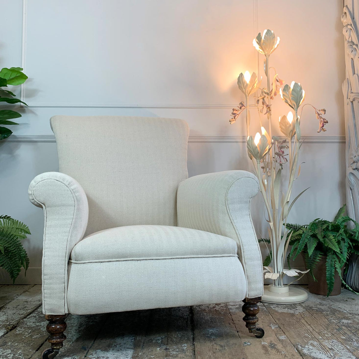 An upholstered antique armchair in an off-white textured fabric, positioned in a styled room setting with a lit decorative floor lamp, potted ferns, and a rustic wooden floor.
