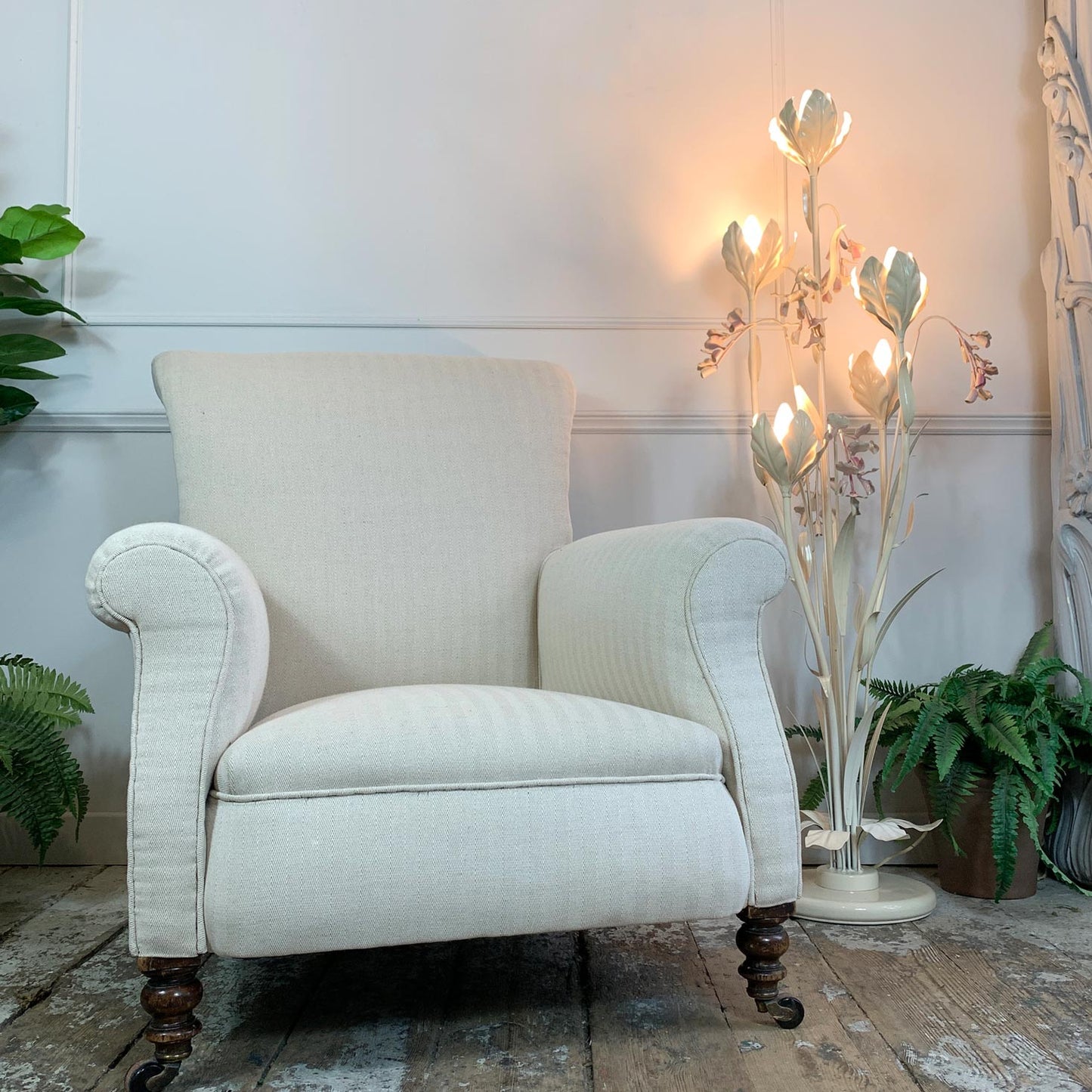 An upholstered antique armchair in an off-white textured fabric, positioned in a styled room setting with a lit decorative floor lamp, potted ferns, and a rustic wooden floor.