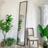A tall, full-length antique tailors floor mirror with a thin black frame leans against a white paneled wall in a styled room. A distressed wooden chair with a light beige upholstered seat sits beside it, surrounded by potted green plants and a large white decorative column.