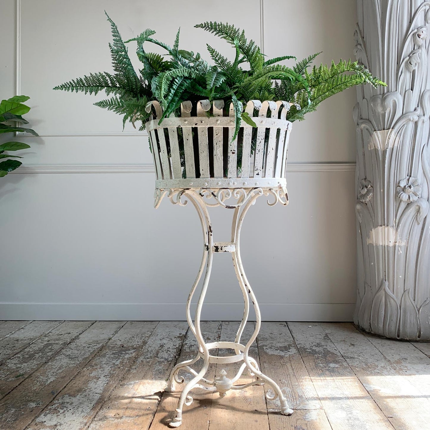 A white metal antique Arras planter basket with a decorative scrollwork stand, filled with lush green ferns, set against a white paneled wall and rustic wooden floor.
