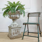 A distressed antique white terracotta urn planter filled with green ferns, placed beside a teal-green metal chair on a beige carpet against a white paneled wall.