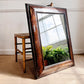 A large rectangular William and Mary wall mirror with an ornate distressed dark brown wooden frame, leaning against a wall next to a bamboo-style stool on a beige carpet.