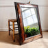 A large rectangular William and Mary wall mirror with an ornate distressed dark brown wooden frame, leaning against a wall next to a bamboo-style stool on a beige carpet.