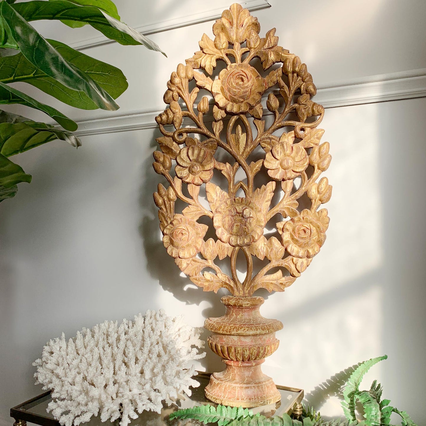 An ornate carved wooden decorative altar vase and palm featuring a floral tree design on a pedestal base, styled on a tabletop with a white coral piece and green plants against a white wall.